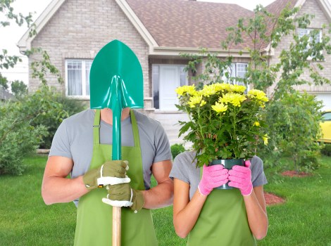 A gardener and a woman are standing in a neatly maintained front garden with a modern brick house in the background. The gardener, dressed in a grey t-shirt, green apron, and yellow gloves, holds a small green lawn spade with its blade covered by a green garden cover. The woman, also wearing a green apron and bright pink gloves, is holding a large pink pot of vibrant yellow flowers, possibly chrysanthemums or potted daisies. The garden features a lush, well-trimmed lawn with dense green shrubbery bordering the area, and young trees with fresh green leaves. The house has light-colored brick walls, white framed windows, and a grey tiled roof, suggesting a typical residential property in Hayes. The scene is captured under natural daylight with clear weather, indicating a pleasant outdoor environment suitable for gardening and lawn care activities. This image reflects professional gardening practices and outdoor maintenance, aligning with services offered by Gardening Services Hayes in the Hayes area, postcode HA9, enhancing outdoor aesthetics with floral displays and lawn care.